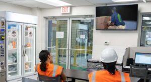 Two factory workers sitting in breakroom, watching TV of Alchemy looping video about back safety