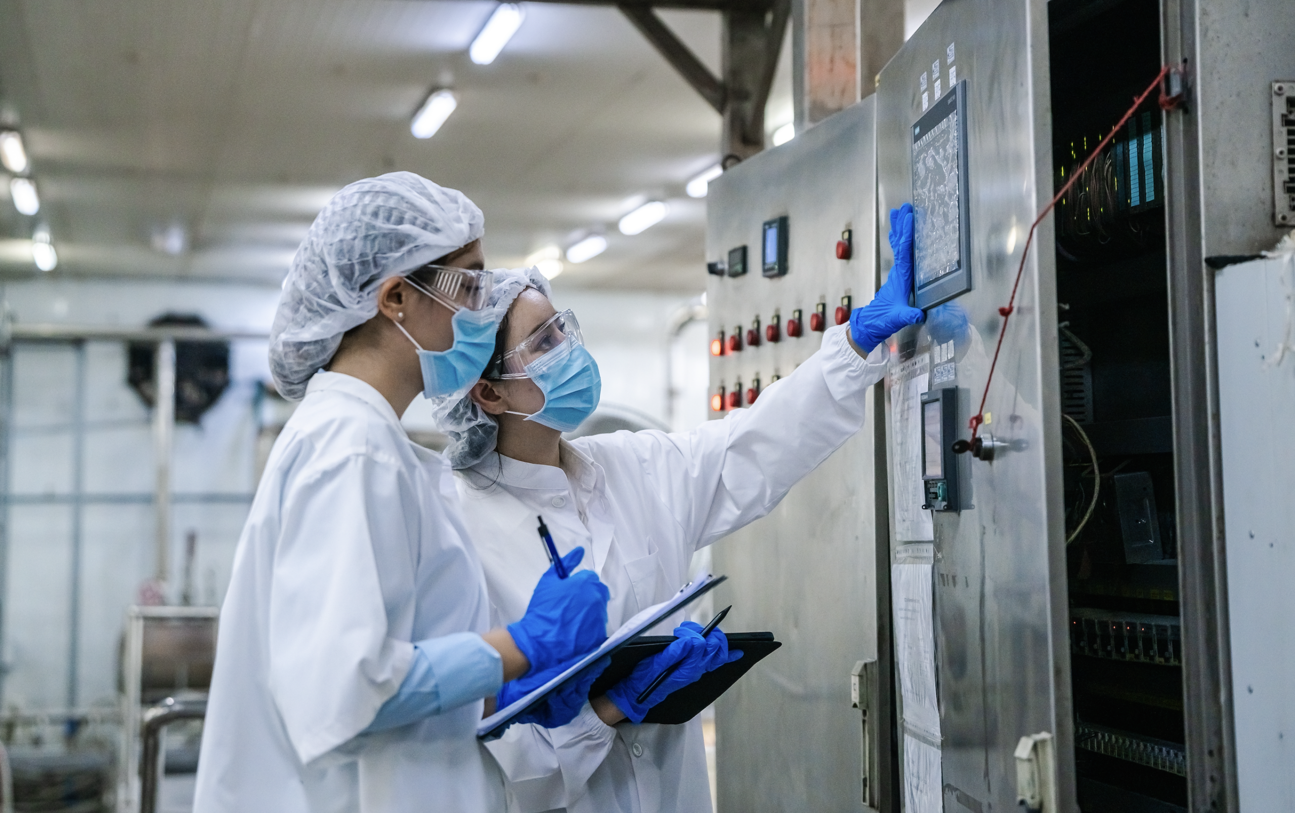 Two food plant workers in hairnets, safety glasses, masks, and gloves check a machine control panel while one records observations on a clipboard