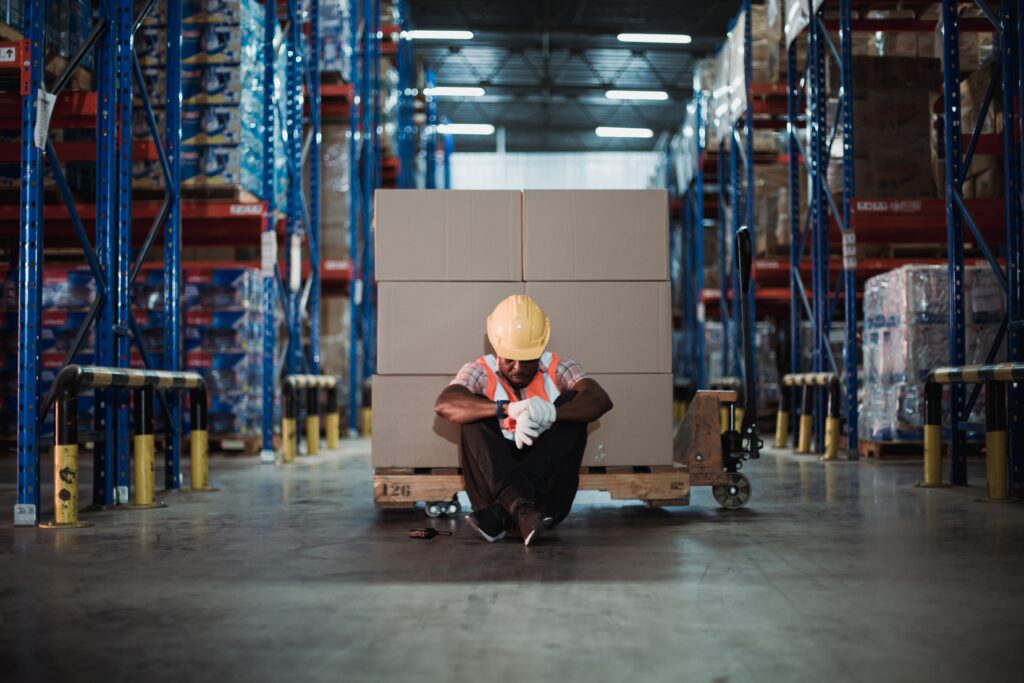 Warehouse worker in a hard hat and safety vest sits against a pallet between storage racks, head bowed, showing the strain when growth outpaces know-how